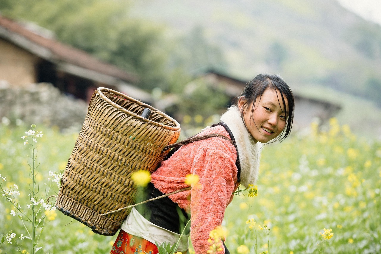 A woman in a flowered field carries a tall open-weave basket on her back.