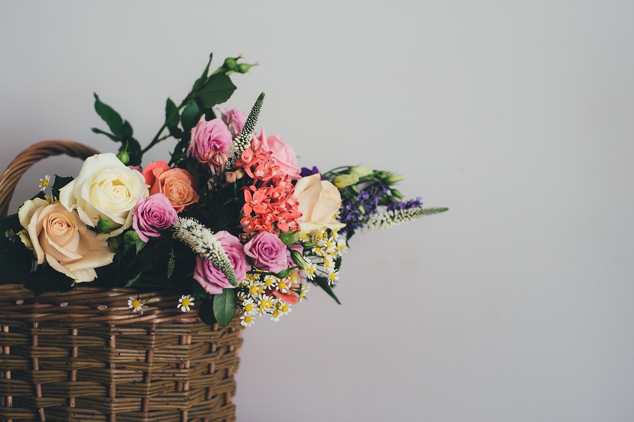 A dark rectangular wicker basket holds a loose arrangement of roses, greenery, and small flowers.