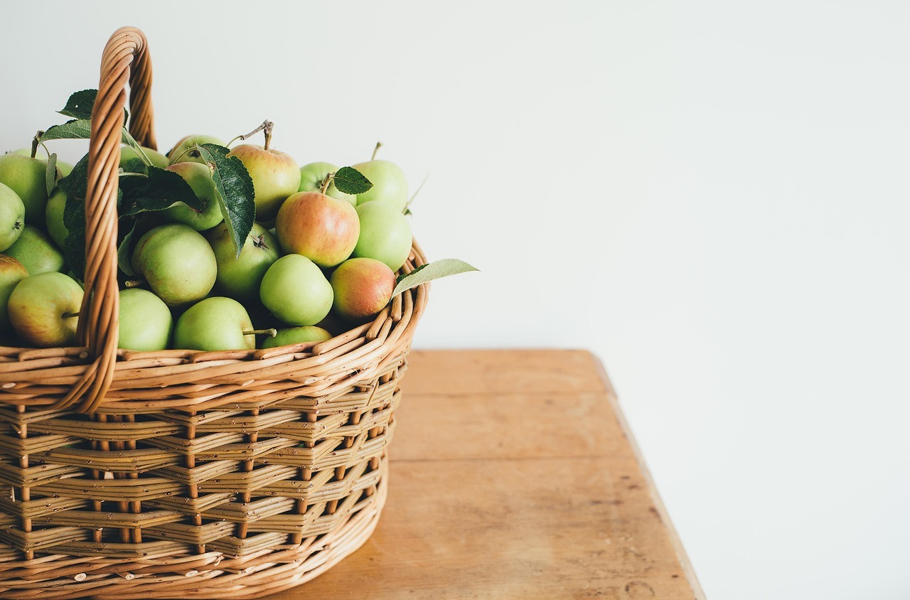 A large wicker basket filled with green and blushed apples sits at the edge of a wooden table.
